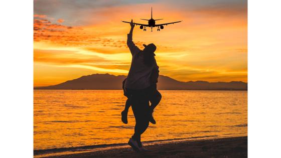 Mann bærer mann på ryggen på en strand i solnedgang med et fly på himmelen.